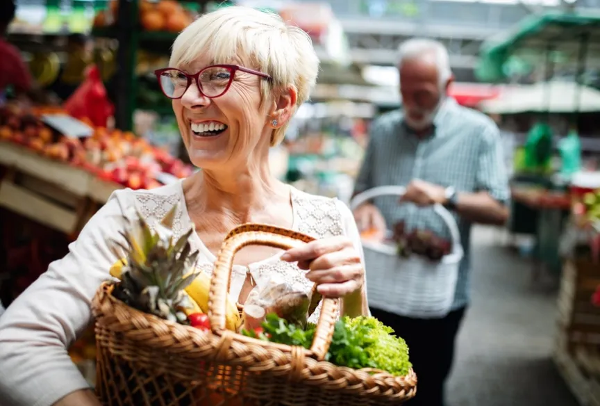 Older smiling woman holding a basket of fruit and vegetables at a market